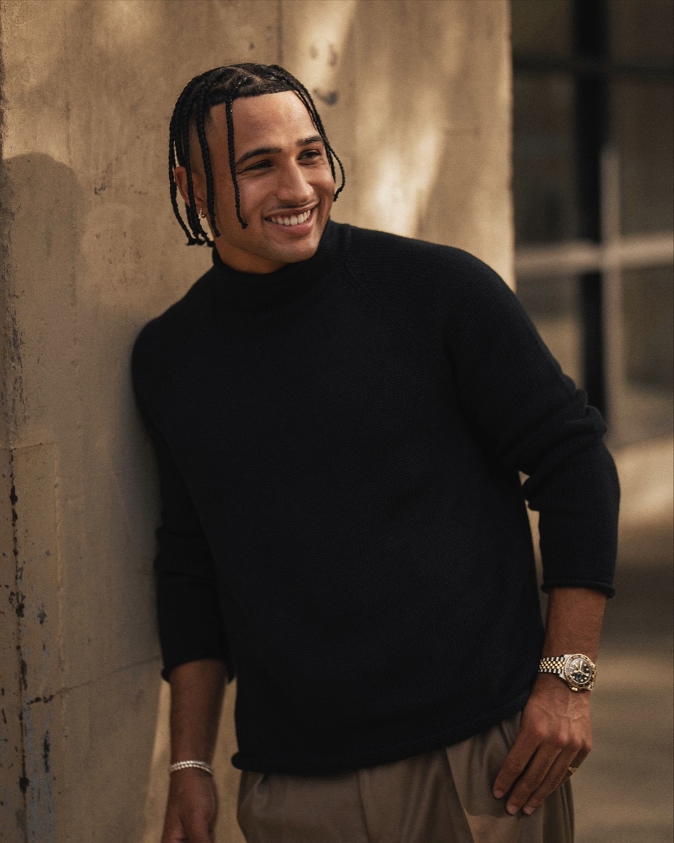 A man in a black rollneck sweater and khaki trousers leaning against a stone wall, laughing, in warm evening light