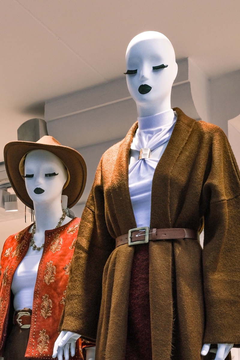 Two store mannequins dressed for autumn, one in a belted brown wool wrap coat and one in a red embroidered suede waistcoat and tan hat