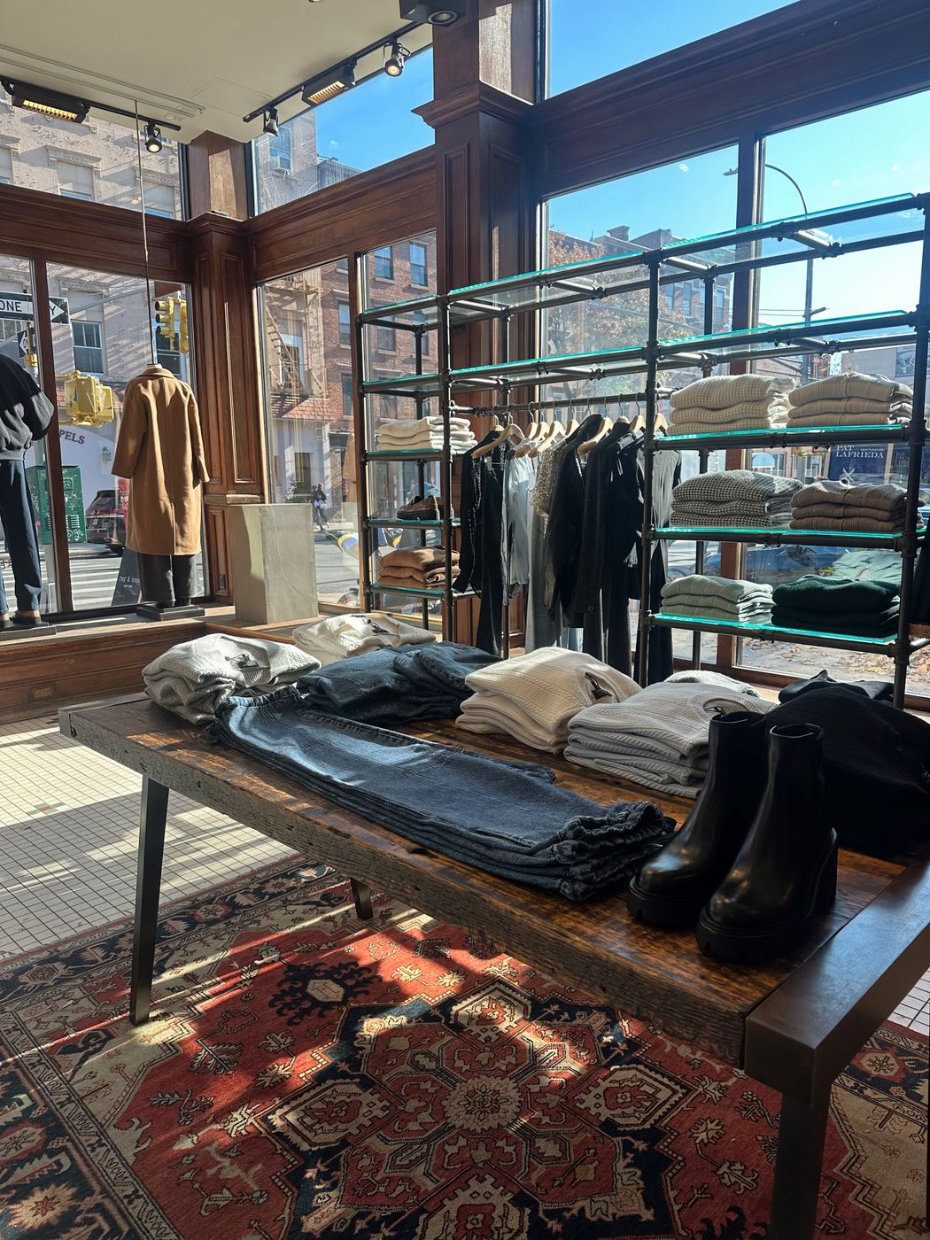 Sunlit boutique interior in New York with folded knitwear and denim on a wooden table, black boots at the end, and a camel coat in the window behind