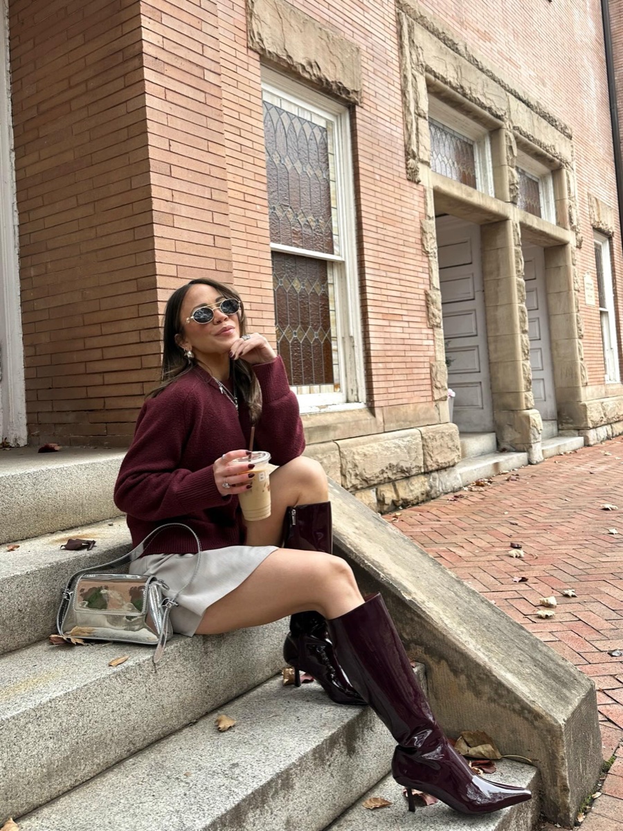 A woman in an oversized burgundy knit, grey mini skirt and burgundy patent knee-high boots sits on the stone steps of a brick building holding an iced coffee