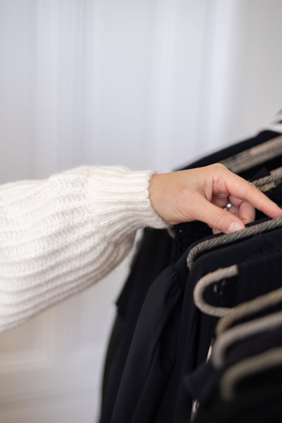 Arm in a chunky cream knit sweater reaching into a rack of dark garments on rope hangers