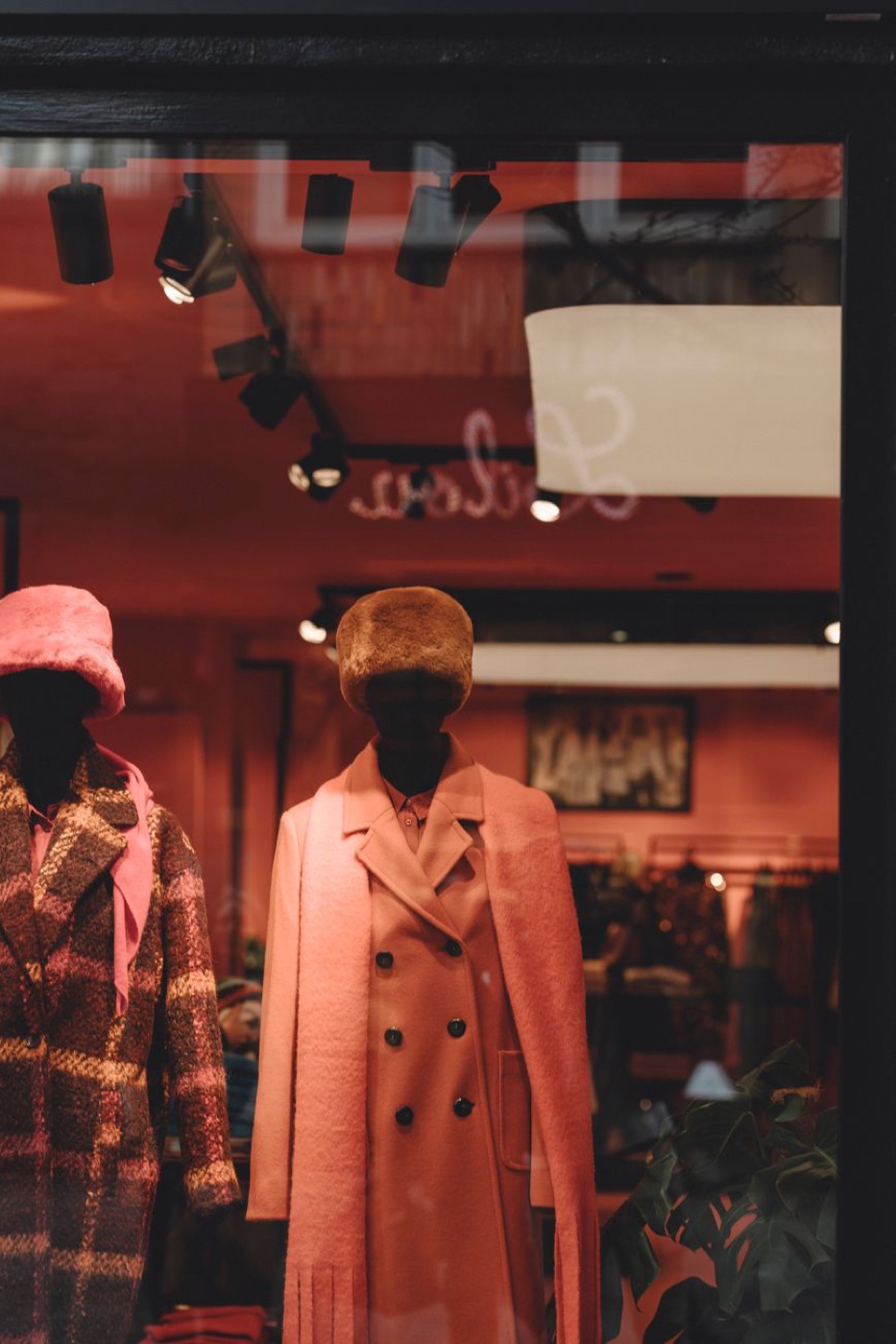 Two mannequins in wool fall coats and faux fur hats behind a shop window with ceiling spotlights reflected in the glass