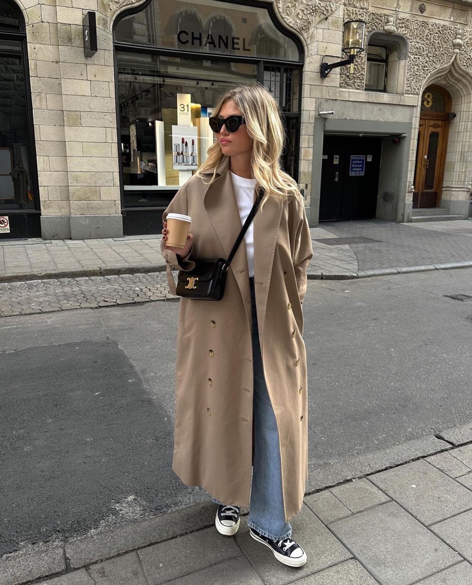 A long camel trench coat worn open over a white tee and jeans with black sneakers, carrying a coffee, on a stone pavement outside a Chanel boutique
