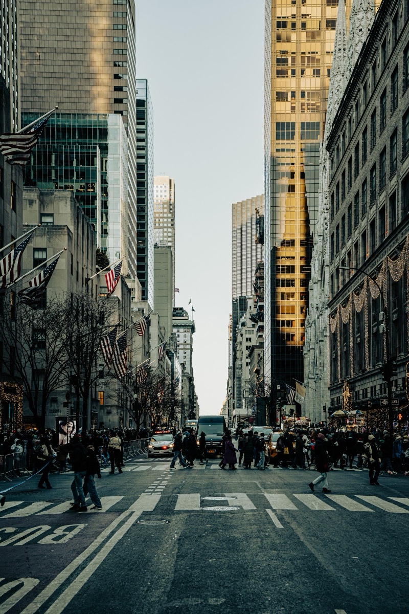 Fifth Avenue in New York looking north, American flags on the left buildings, crowds crossing at the light between tall glass towers