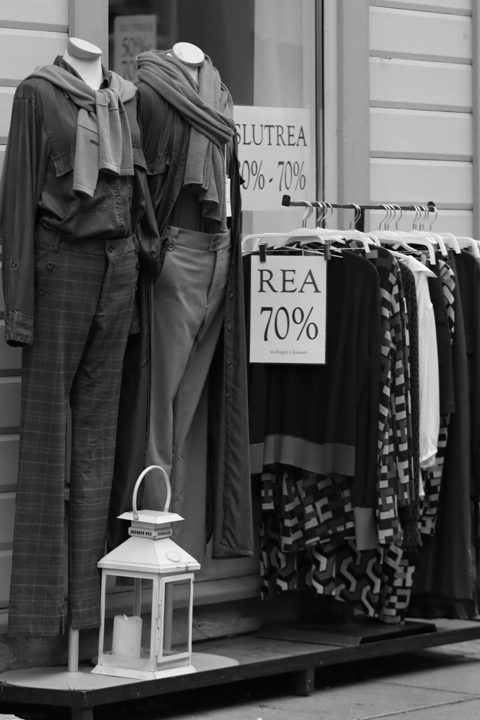 Black and white photo of a storefront with mannequins and a clothing rack under signs reading SLUTREA and REA 70%
