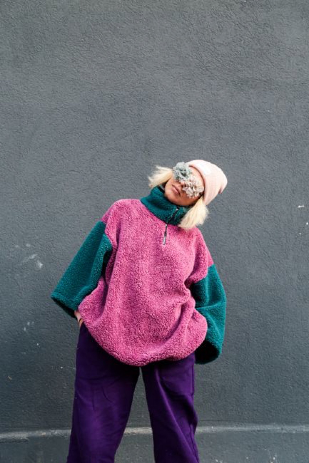 A person in an oversized pink and teal colorblock pile fleece pullover and pink beanie leaning against a gray wall, face hidden behind a sprig of dried flowers