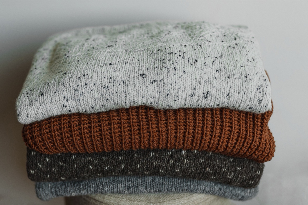 A stack of four folded knit sweaters in speckled white, rust, charcoal, and grey on a linen surface