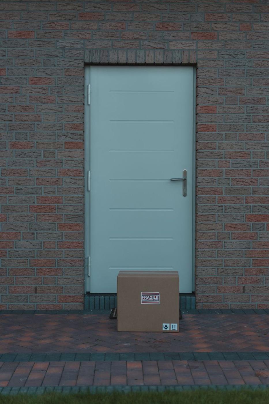 A single brown cardboard box with a FRAGILE sticker sits against a pale green door in a brick wall