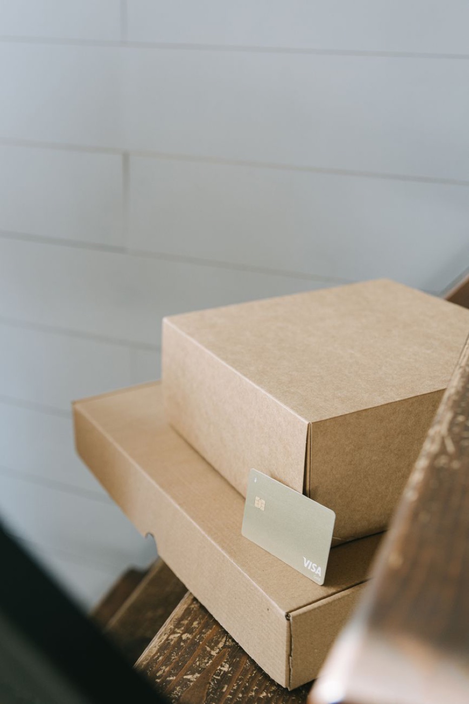 A plain gold Visa card leans against two stacked kraft shipping boxes on a wooden stair by a white plank wall