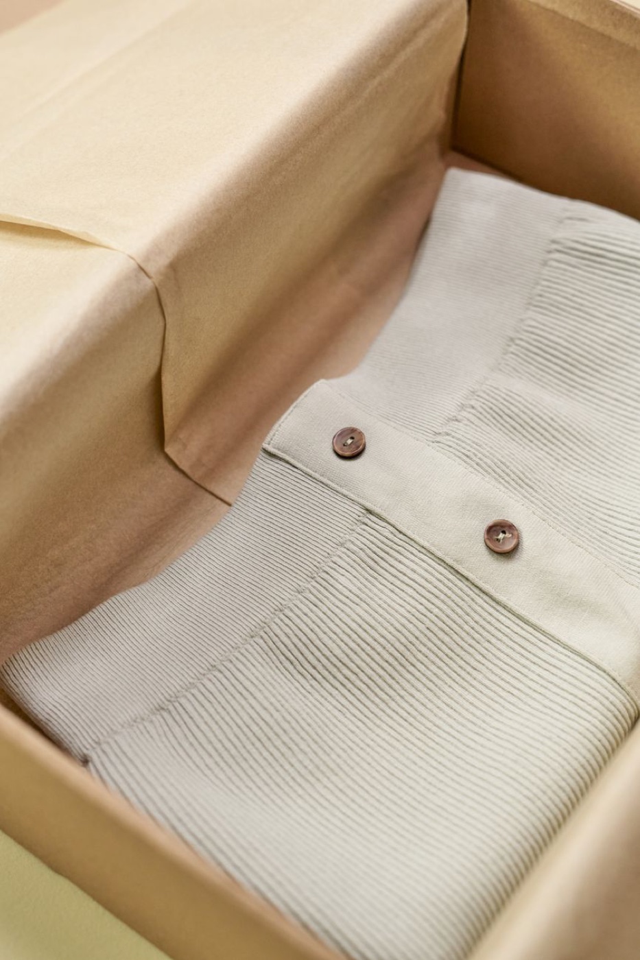 A folded sage green ribbed knit garment with two brown buttons sits in a kraft box lined with tissue paper