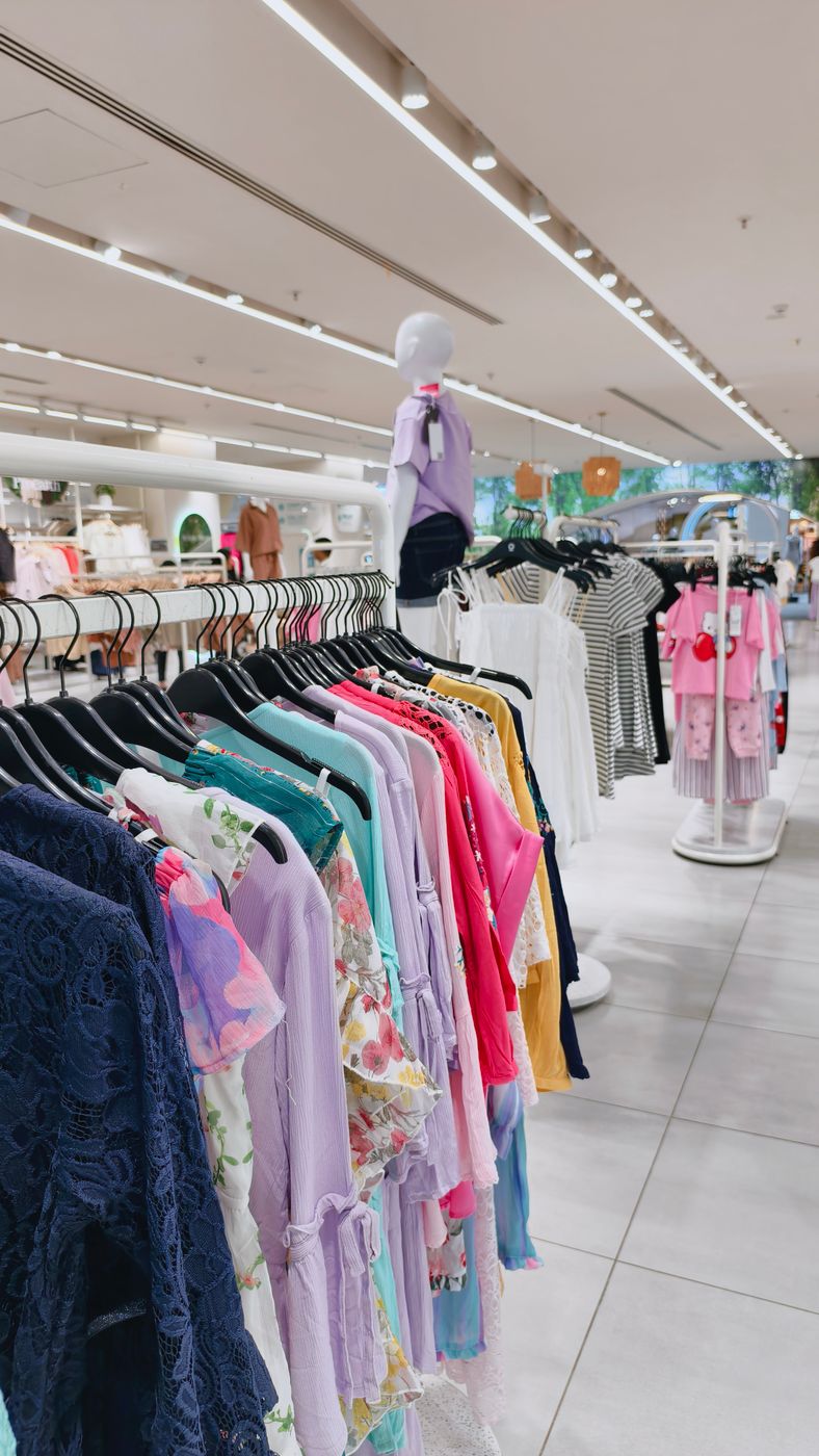 A long rail of colourful blouses and dresses on black hangers in a brightly lit department store, mannequins and further rails behind