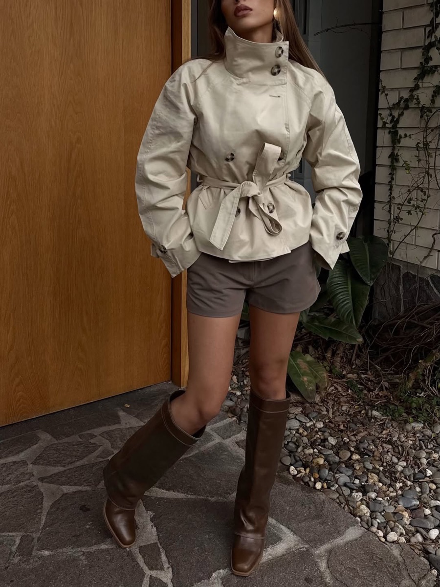 Woman in a cropped stone cotton jacket with a tall buttoned funnel collar and tie belt, brown shorts and knee boots, standing at a wooden door