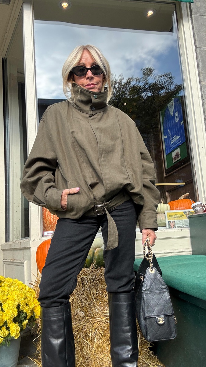 Woman in sunglasses and an olive cotton jacket with a snapped stand-up funnel collar and buckled belt, black jeans and tall boots, outside a shop window with pumpkins
