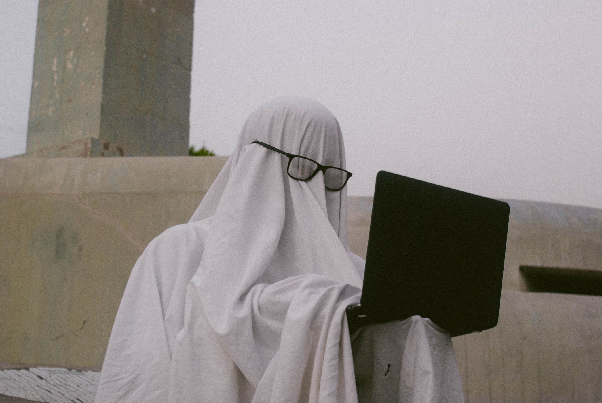 A person draped in a white sheet ghost costume wearing glasses over the fabric while typing on a laptop outdoors