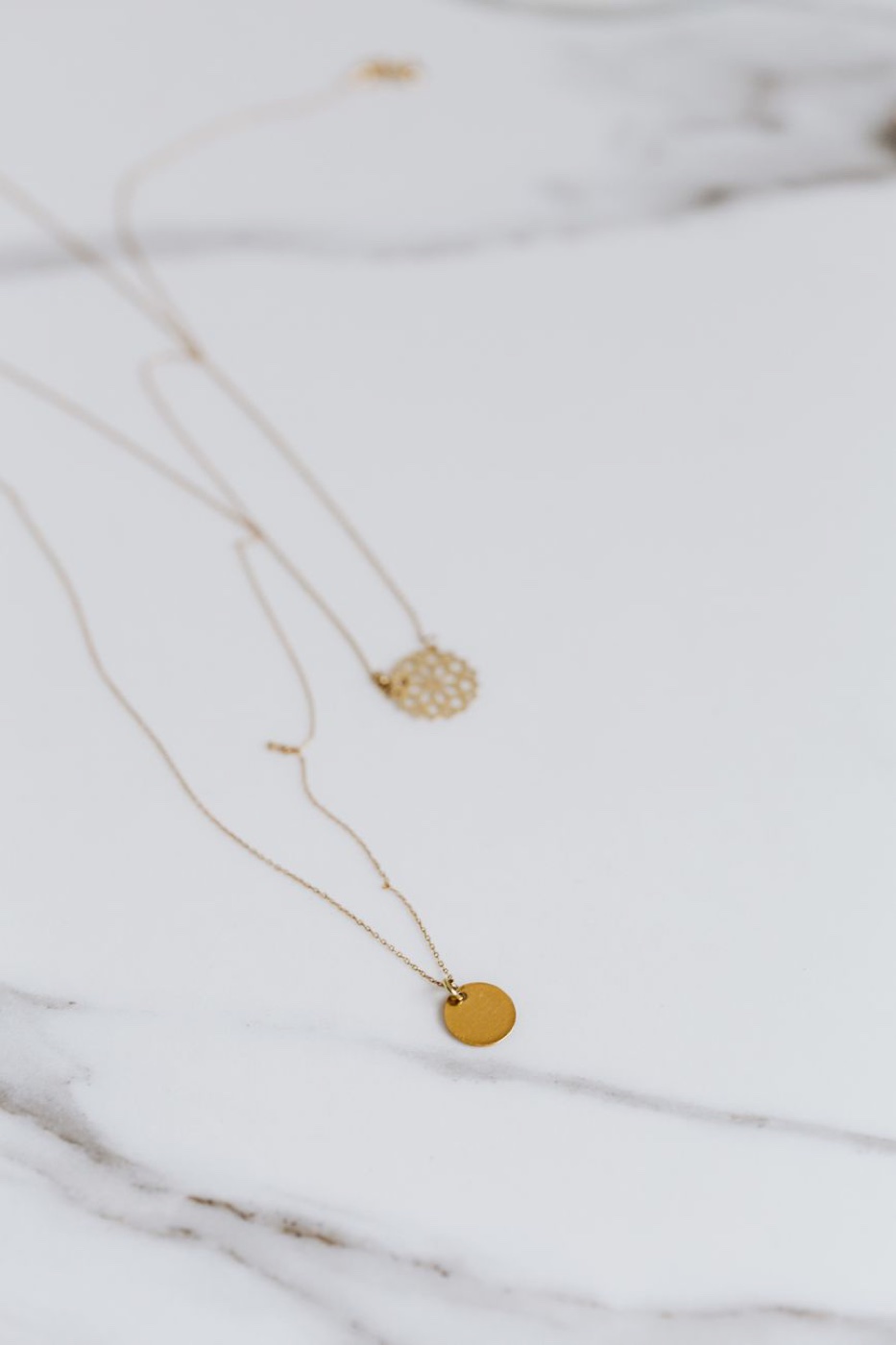 Two fine gold-tone chains with small round pendants laid out on a white marble surface
