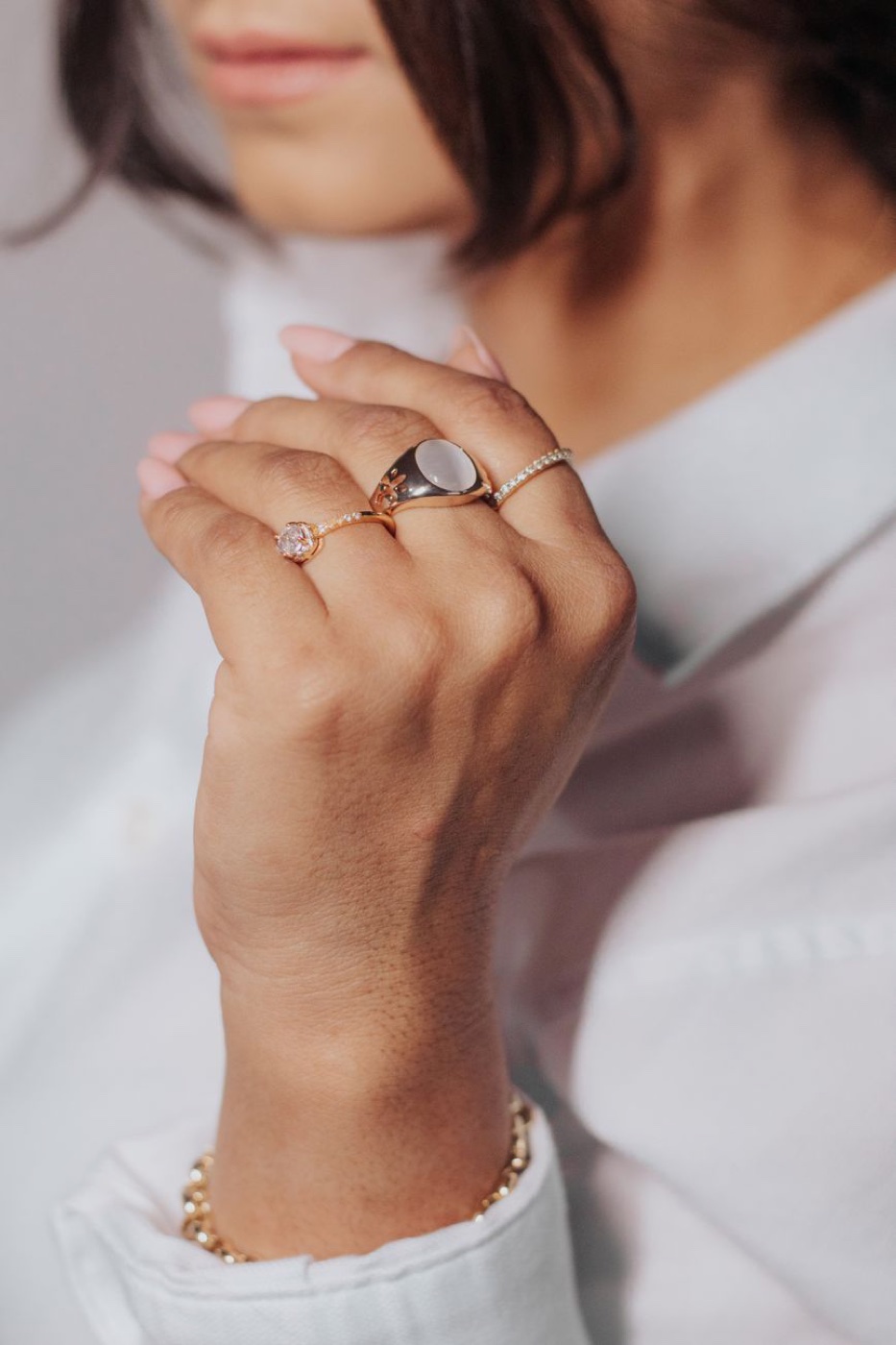 A hand wearing three slim gold-tone rings, a signet among them, resting against a white shirt collar