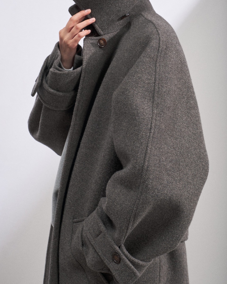 Close detail of a grey melange wool coat with a stand collar, horn buttons and a buttoned sleeve tab, hand at the collar