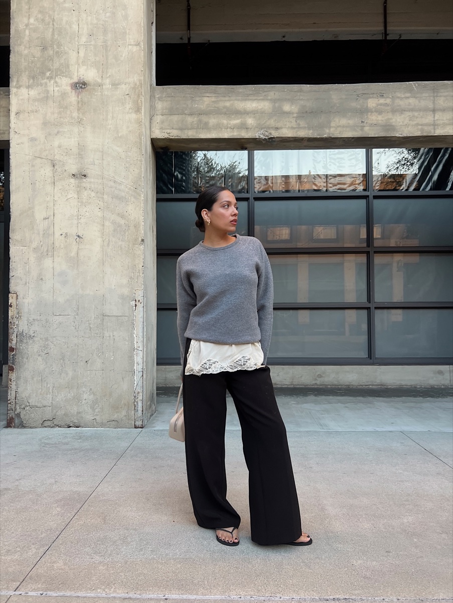 A woman in a grey wool crewneck layered over a lace top with black trousers, standing on concrete outside an industrial building