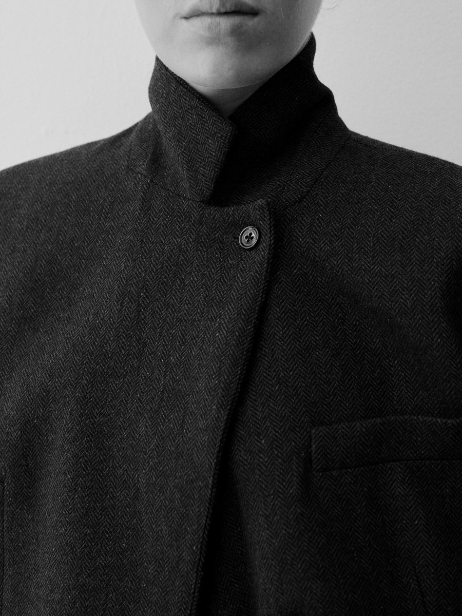 Black and white close-up of a herringbone blazer buttoned to a high standing collar, chin just visible above it