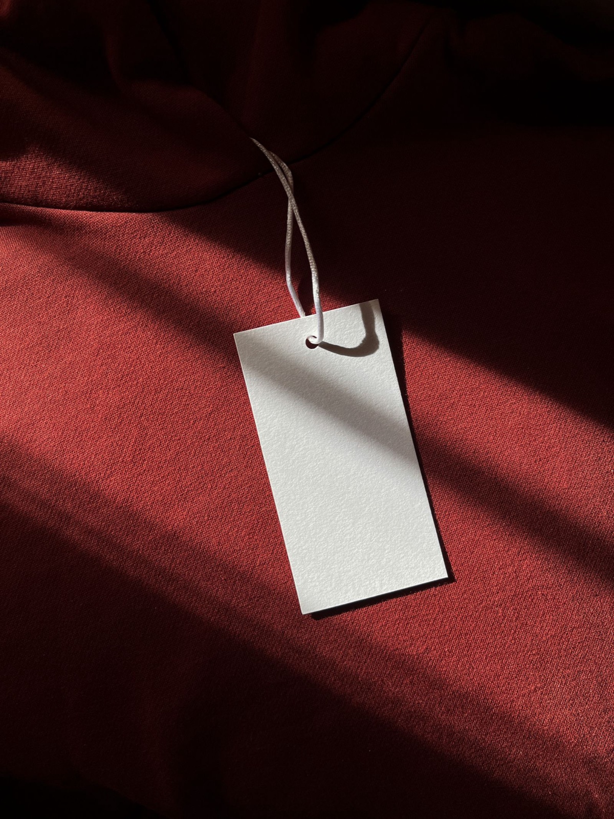 A blank white hang tag on a string resting on rust-red fabric in hard afternoon light
