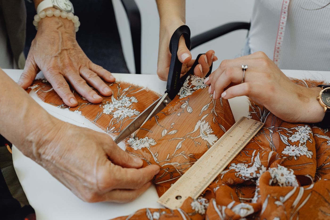 Three pairs of hands at a white table cutting patterned fabric with shears, a wooden ruler and a tape measure in the frame