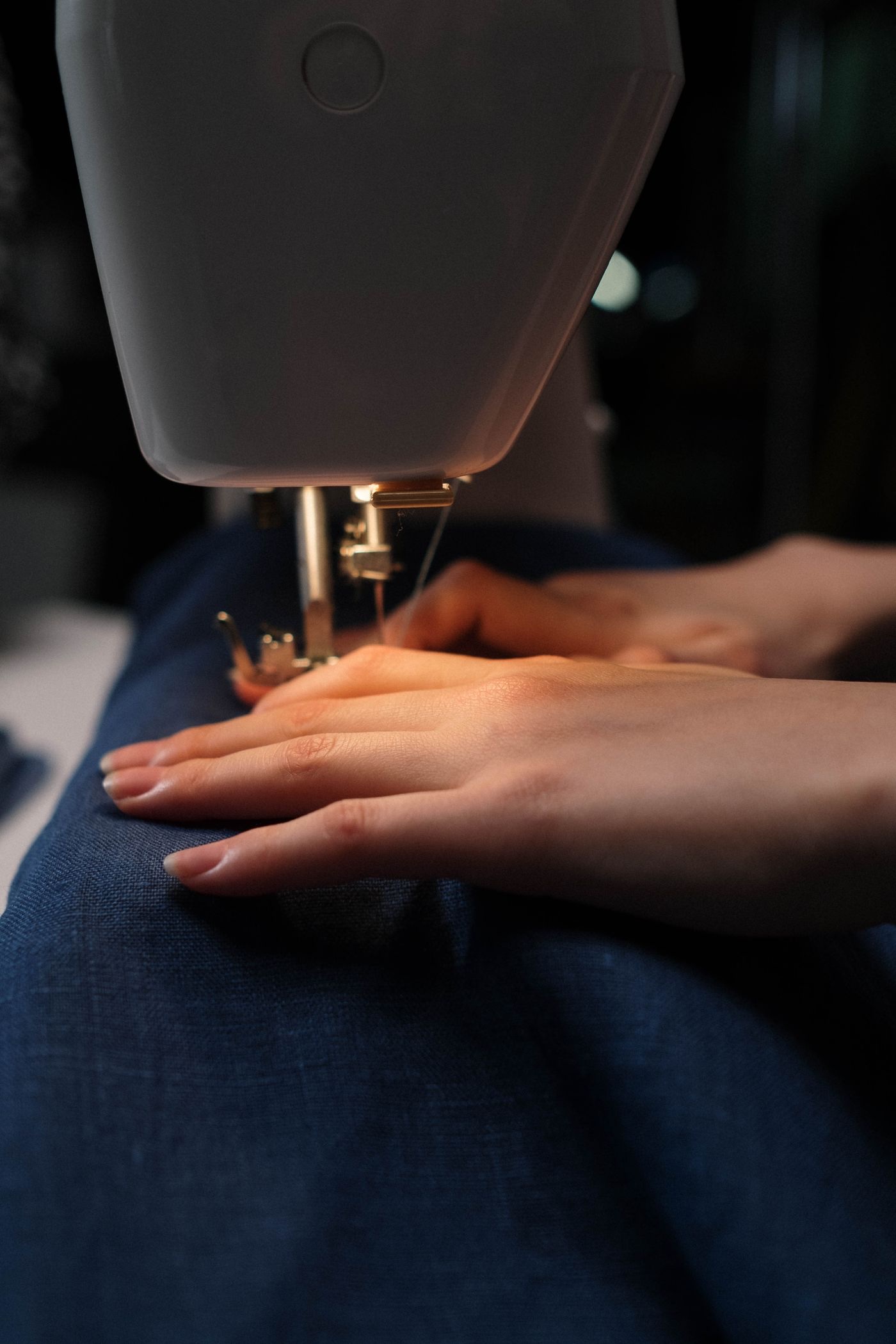 Hands guiding heavy blue linen under the foot of a home sewing machine, lit only by the machine lamp
