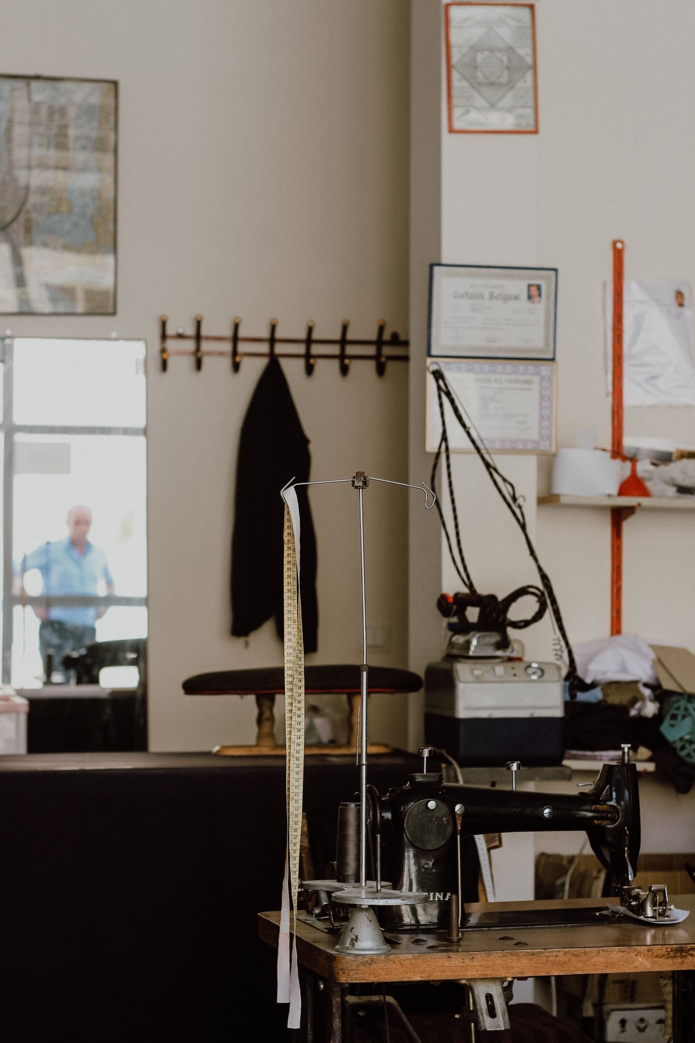 A tailor's workroom with a tape measure hanging from a stand, an industrial sewing machine on a wooden bench and certificates framed on the wall