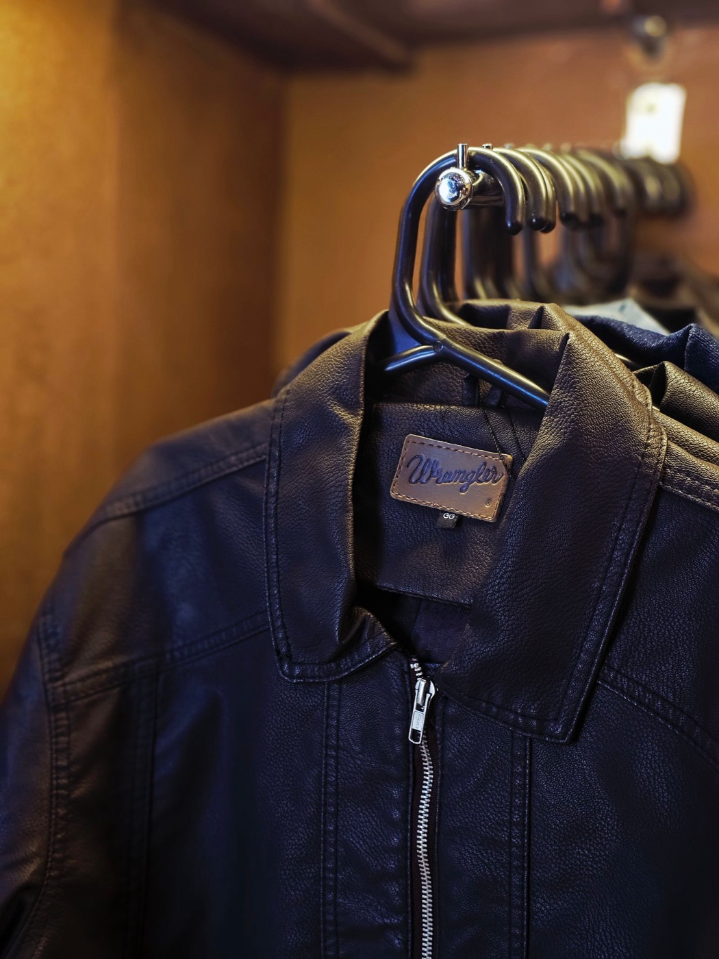 Dark leather jackets hanging on a rack in warm closet light, the front collar turned to show a brown leather brand patch