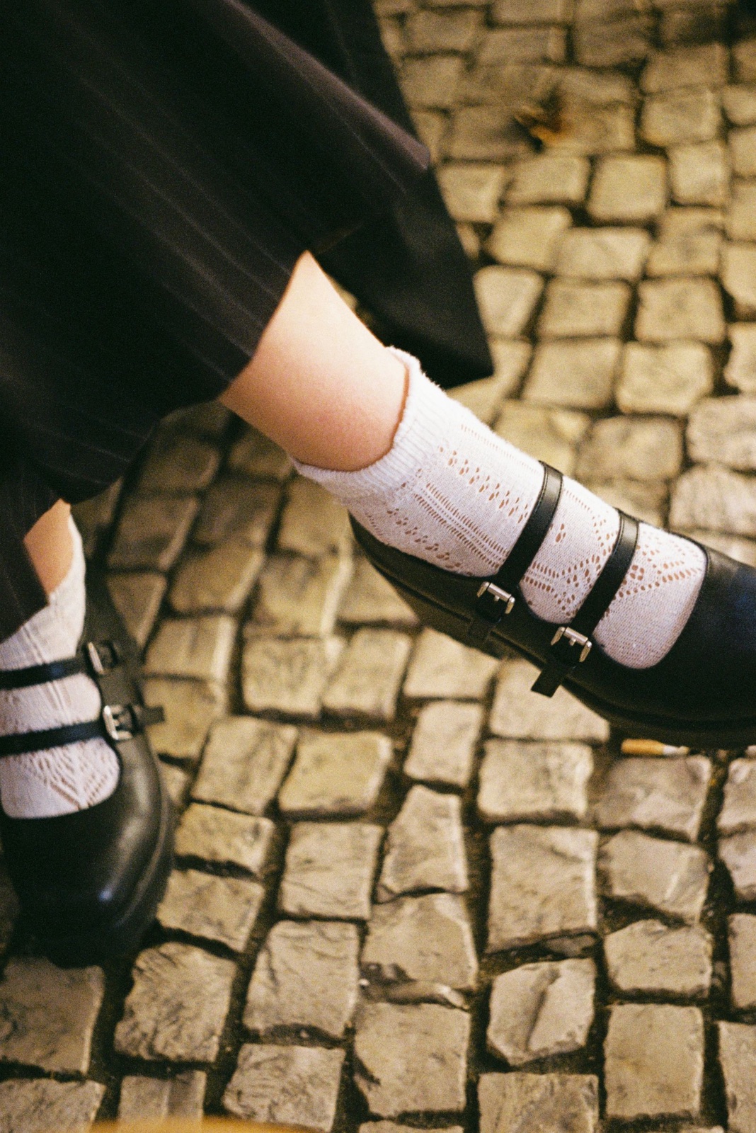 Double-buckle black mary jane shoes worn with white crochet socks and pinstripe trousers on a cobblestone street