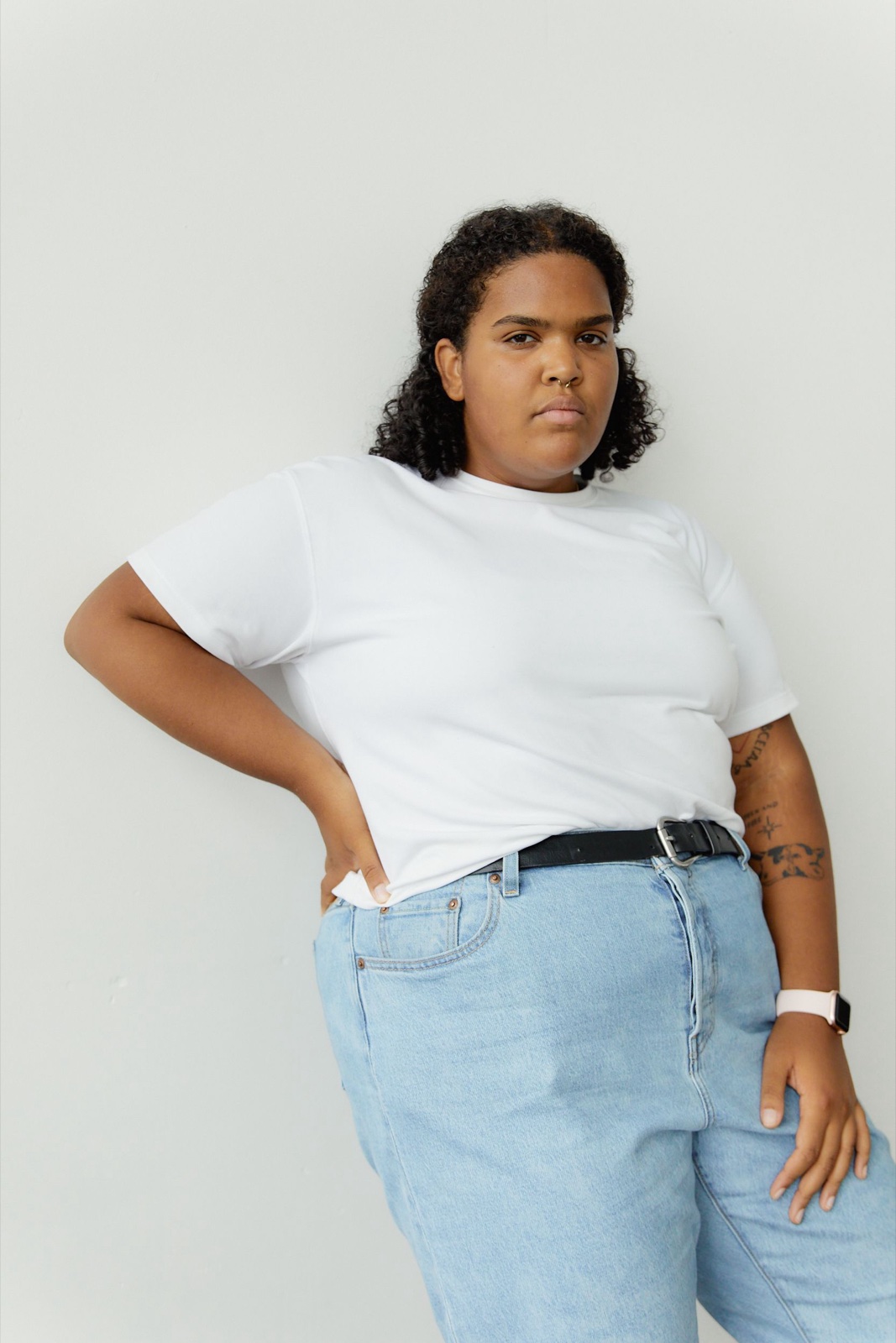 Plus-size woman in a white crew neck t-shirt and light blue jeans standing against a white wall with one hand on her hip