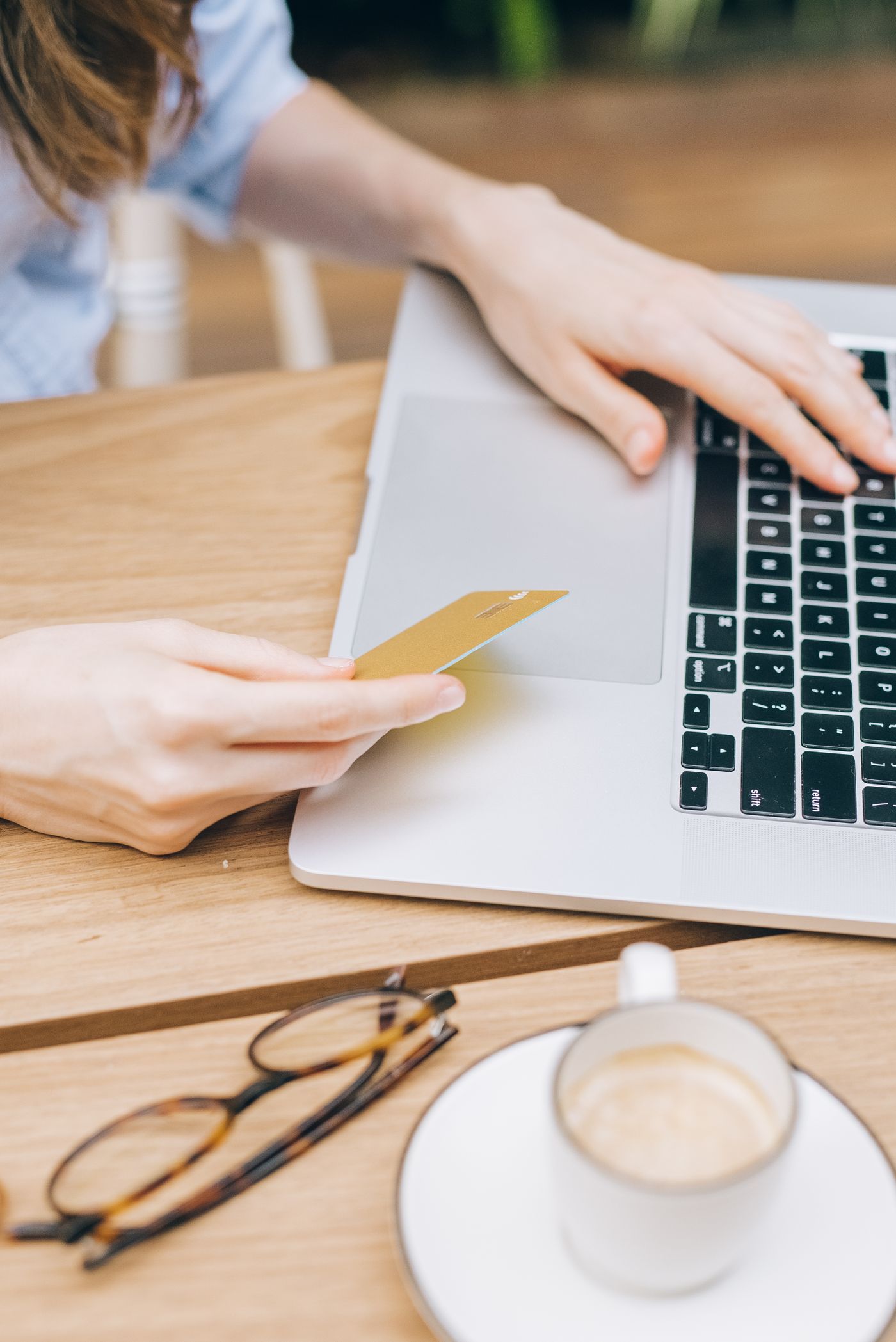 A person at a wooden table holding a gold bank card in one hand while typing on a laptop, coffee and glasses beside them