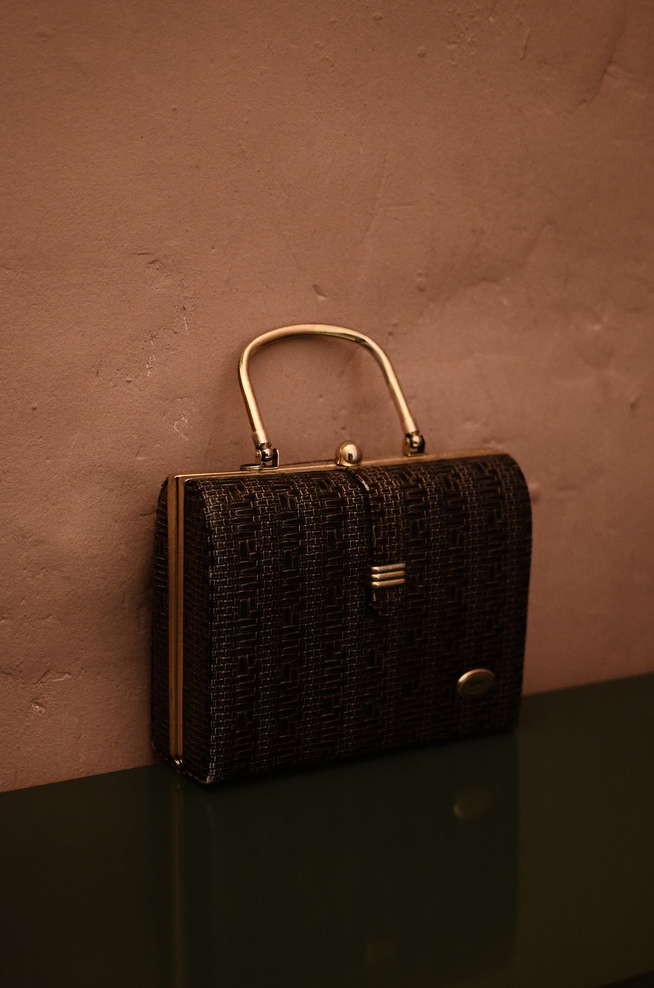 A vintage woven-textured frame handbag with a gold metal handle propped against a rose-toned plaster wall on a dark green shelf