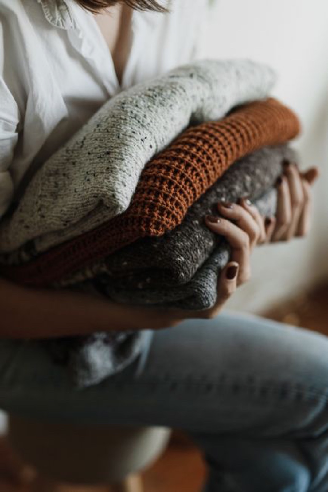 A person in a white shirt holding a tall stack of folded knit sweaters in cream, rust and brown against a plain wall