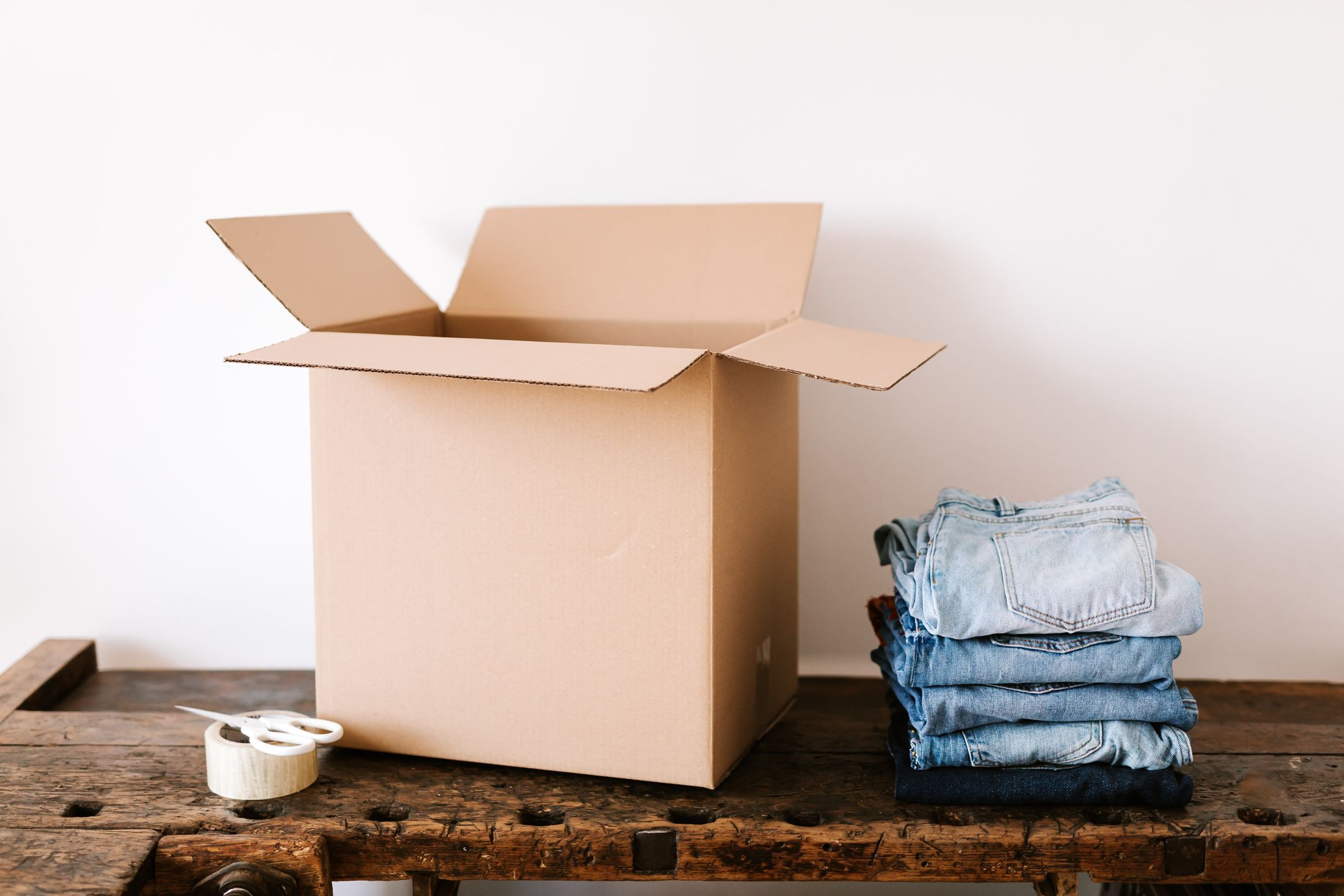 An open empty cardboard box on a wooden bench next to a stack of folded jeans against a white wall