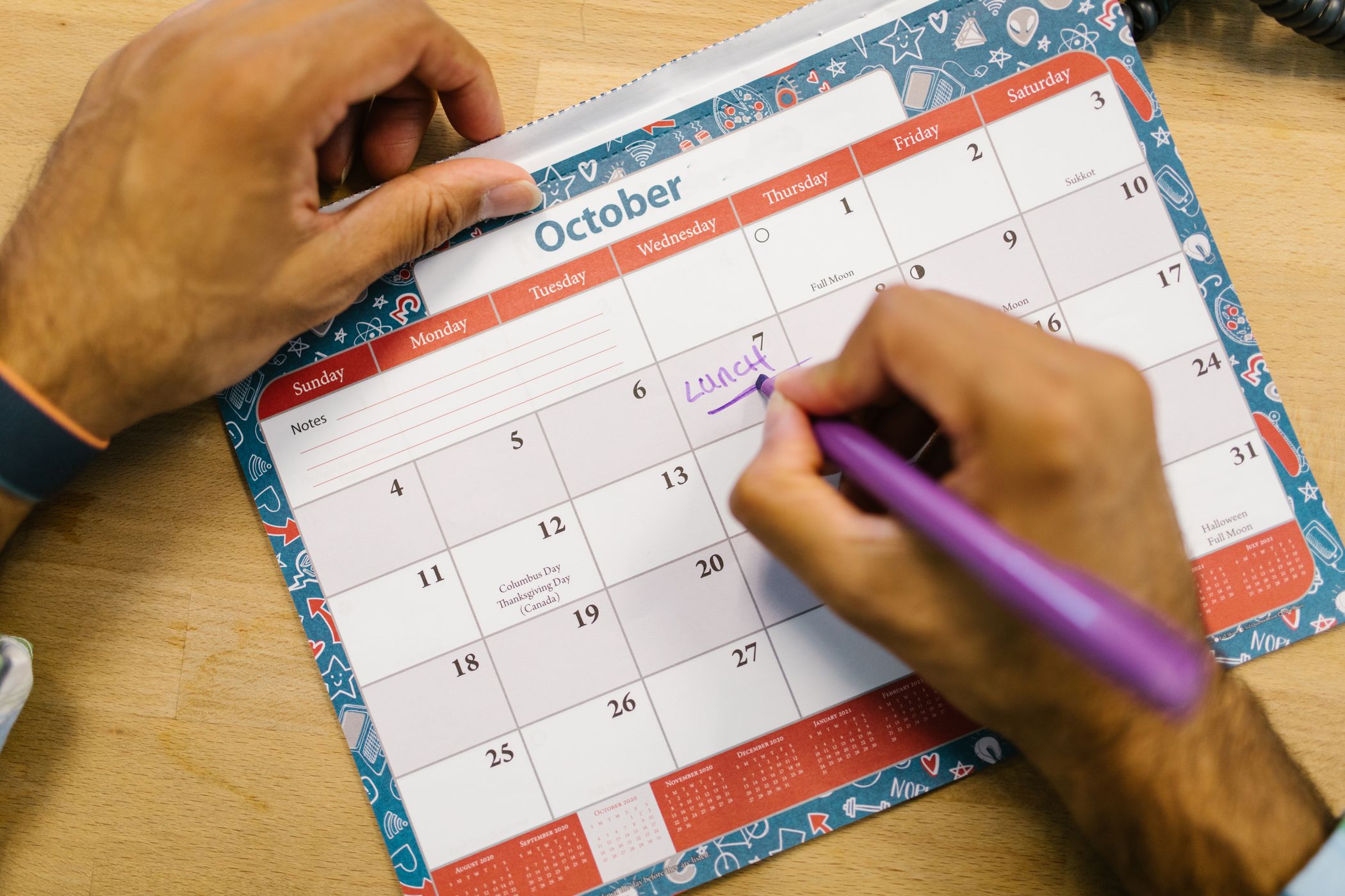 Hands writing a note on a paper desk calendar open to the month of October