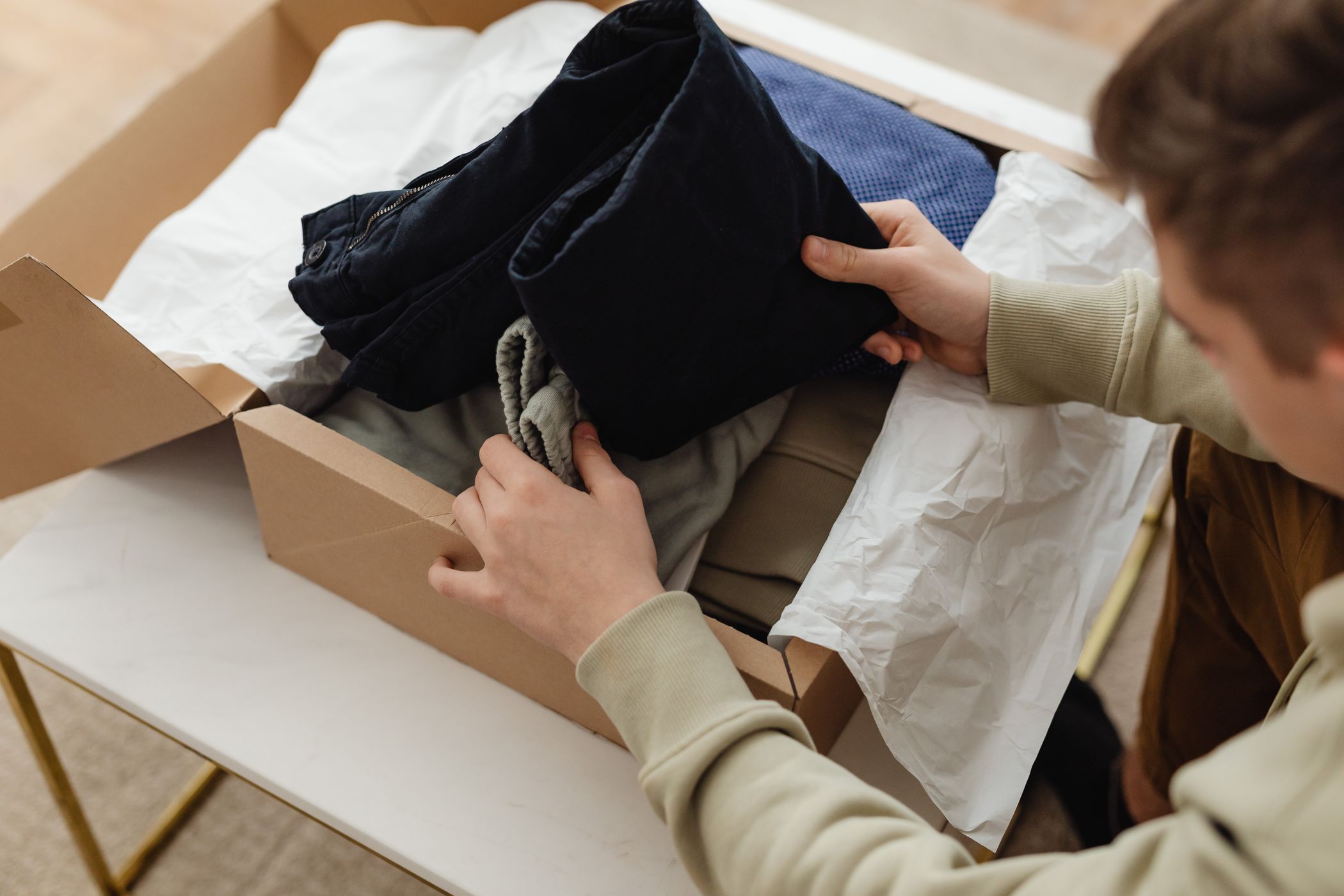 A person lifting folded navy and blue garments out of a cardboard delivery box lined with tissue paper