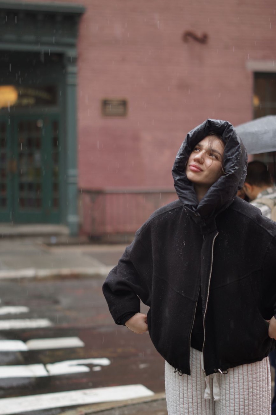 A person in a black hooded jacket standing on a wet New York street in light rain beside a crosswalk