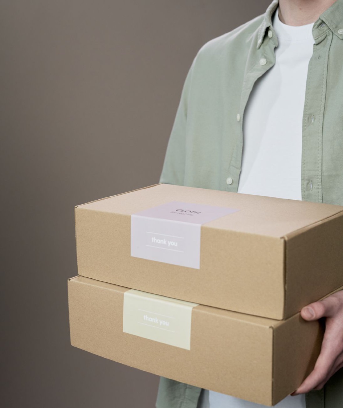 A person in a pale green shirt carrying two stacked cardboard apparel boxes sealed with thank you stickers
