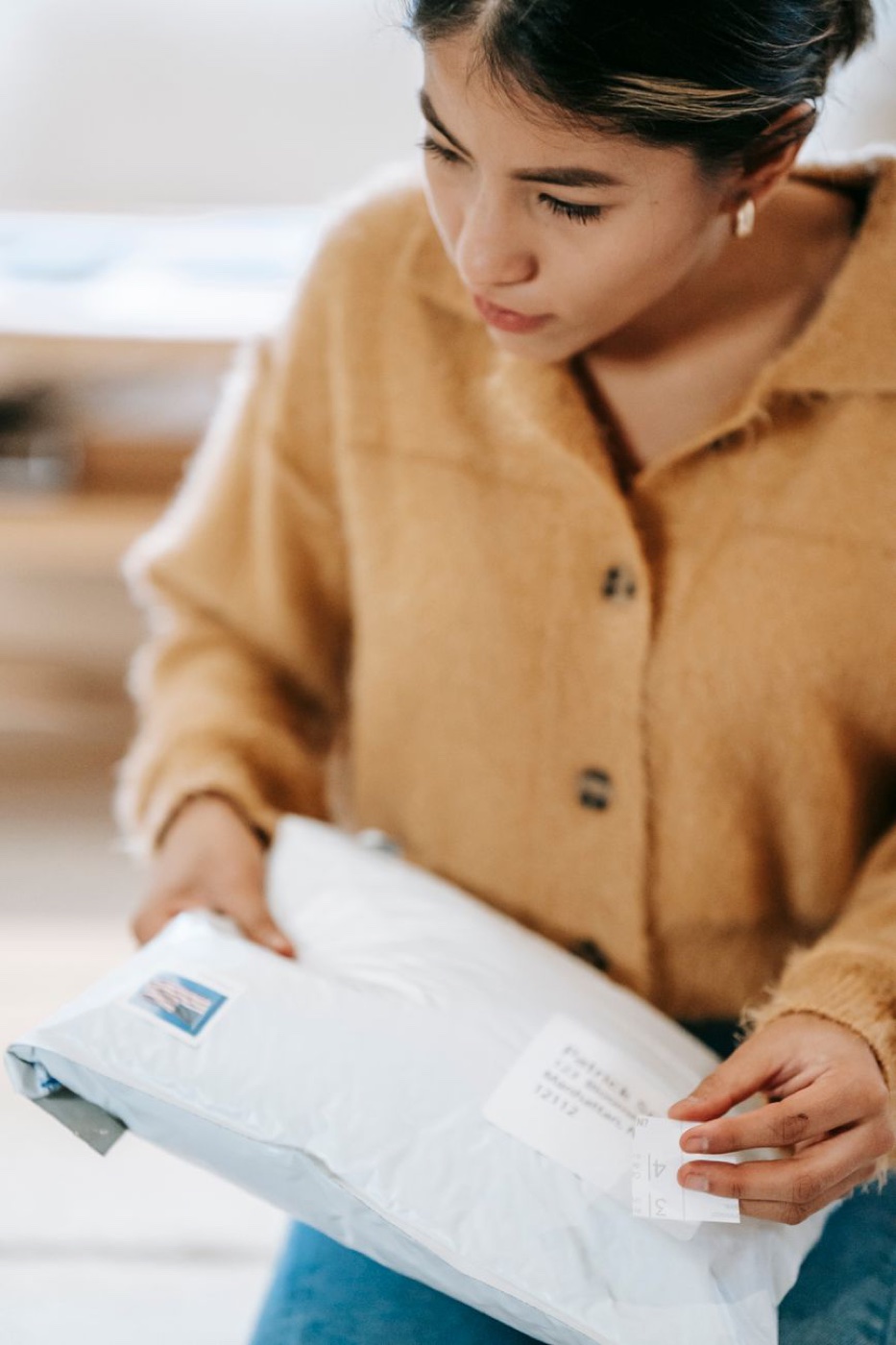 A person in a mustard cardigan holding a white poly mailer and reading the printed address label on the front