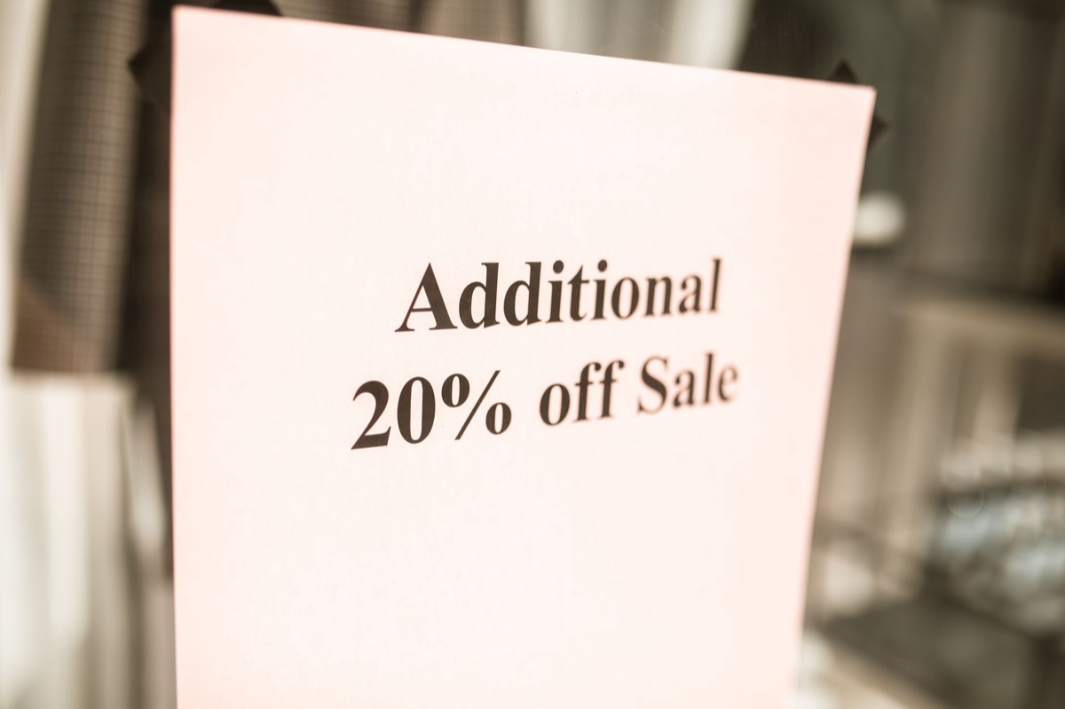 A pale printed card in a shop reading Additional 20 percent off Sale, with a rail of clothing blurred behind it