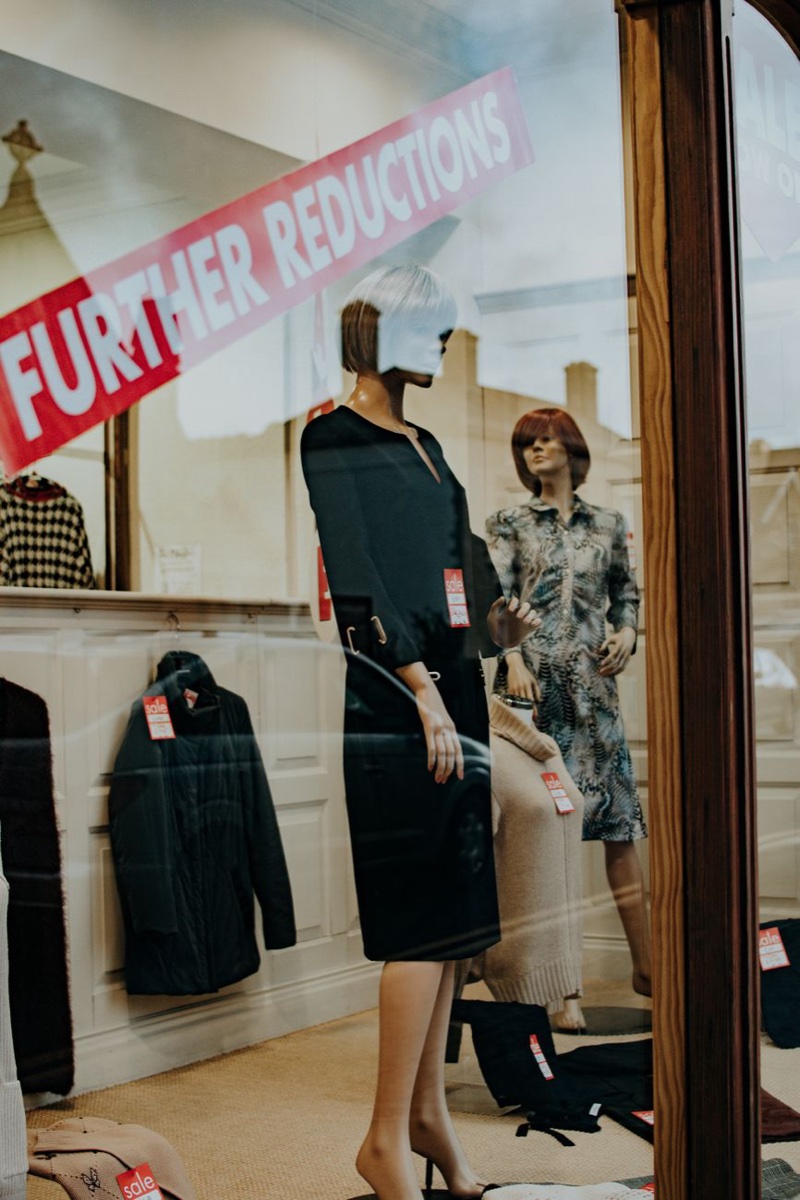 A boutique window with a diagonal red FURTHER REDUCTIONS banner across the glass and small red sale tags pinned to the clothes inside