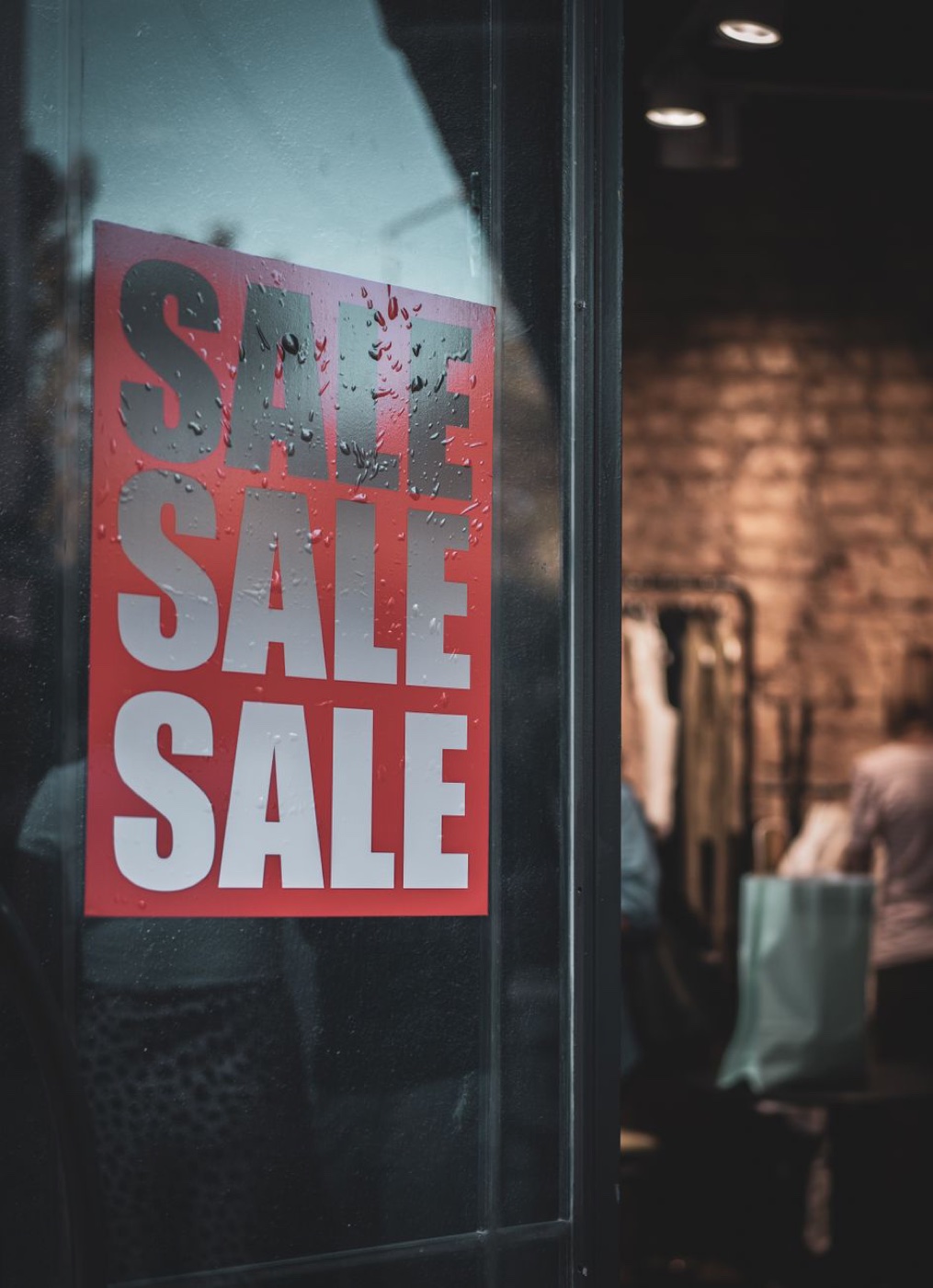 A red poster reading SALE three times taped inside a rain-speckled glass shop door, with a clothing rack and shopper blurred behind it