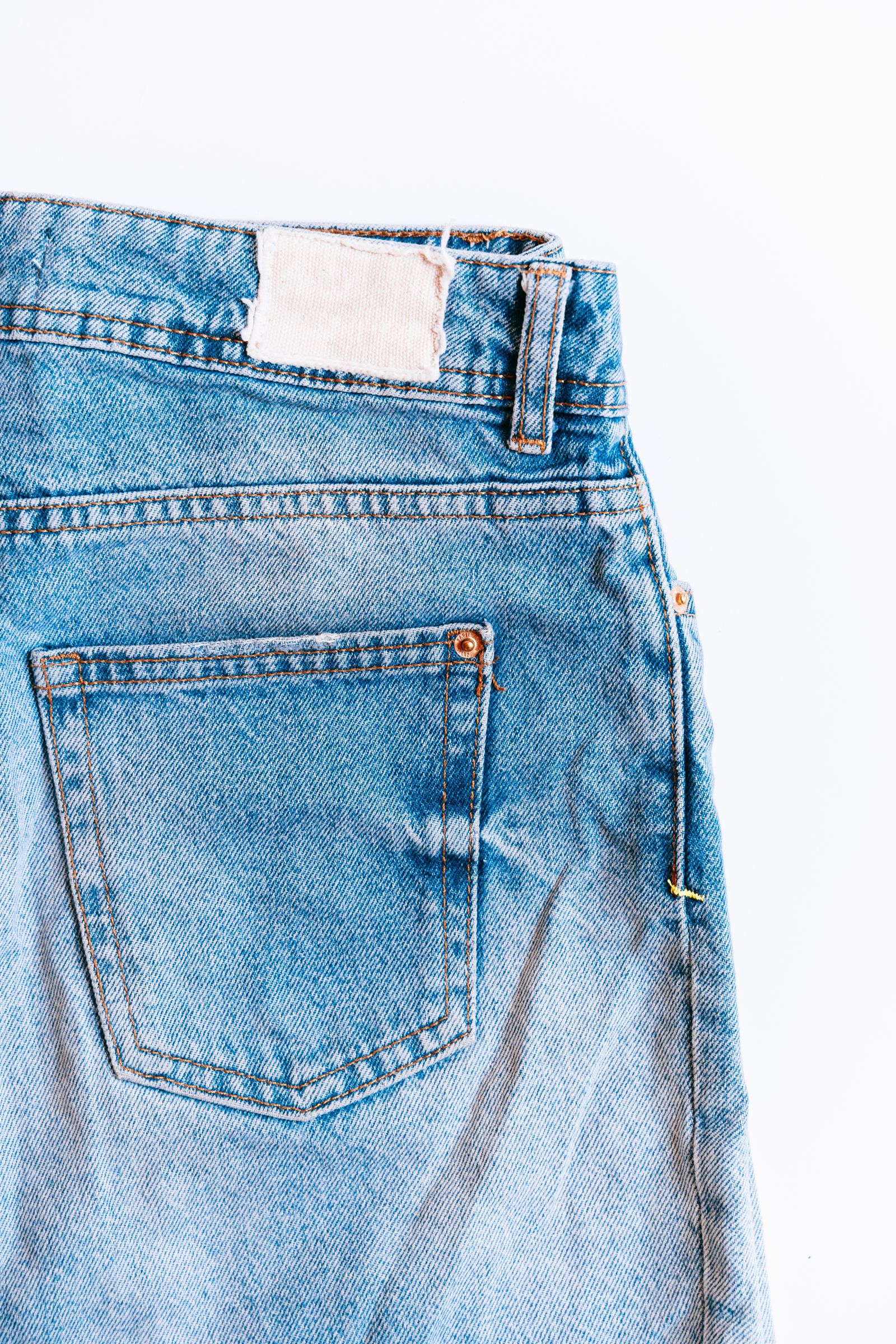 Close-up of a light-wash jean's back waistband with an unprinted beige leather patch and a single belt loop