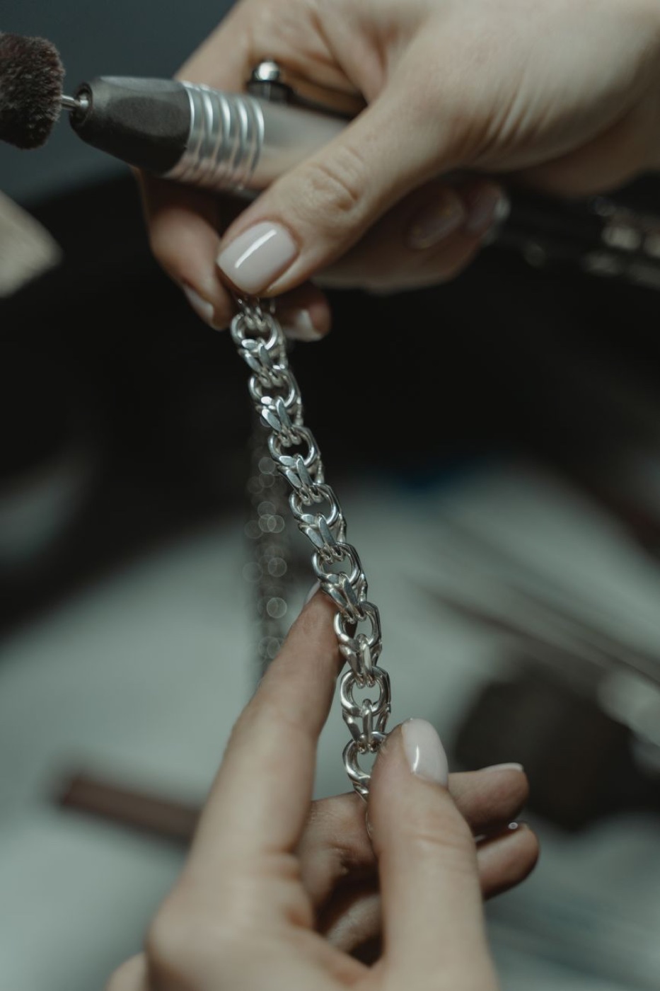 Two hands holding a silver-tone chain against a rotary polishing tool at a jeweller's bench
