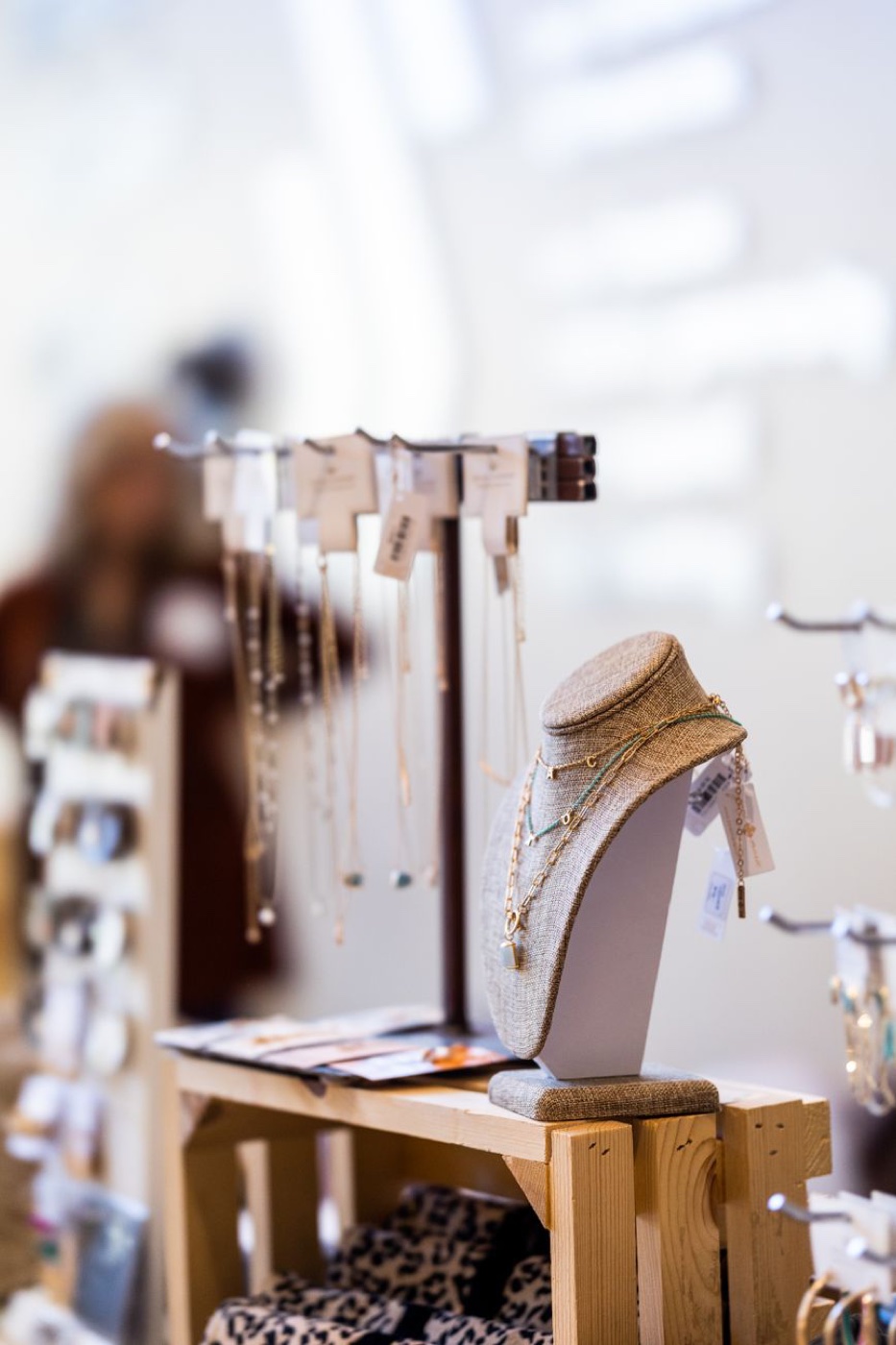 A linen necklace bust and a display rail of chains at a market stall, each piece carrying a small paper hang tag