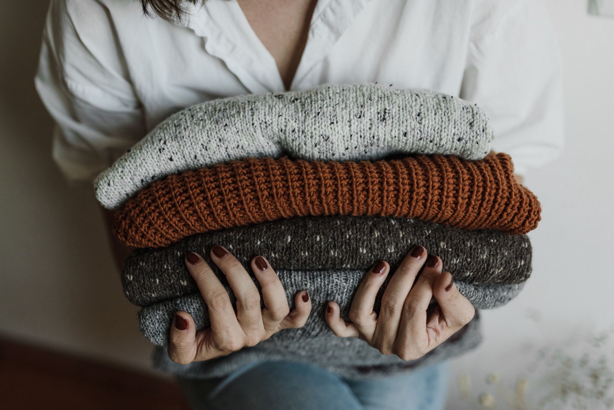 Person in a white shirt holding an armful of folded sweaters in cream, rust and speckled brown