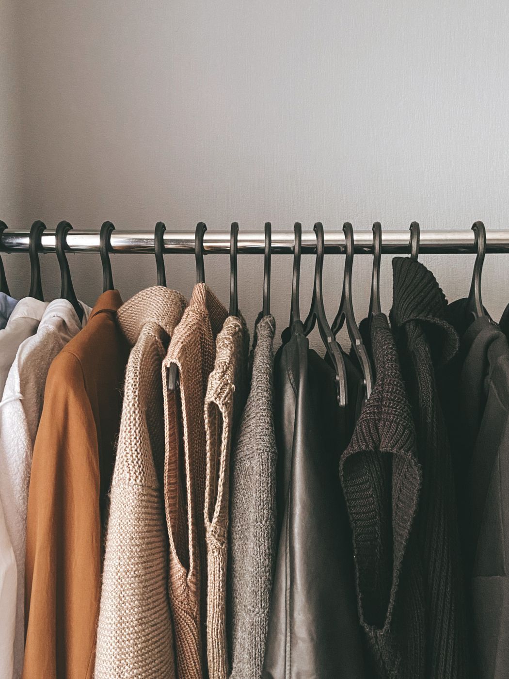 Neutral-toned knits and a rust button-up hanging close together on a closet rod against a gray wall