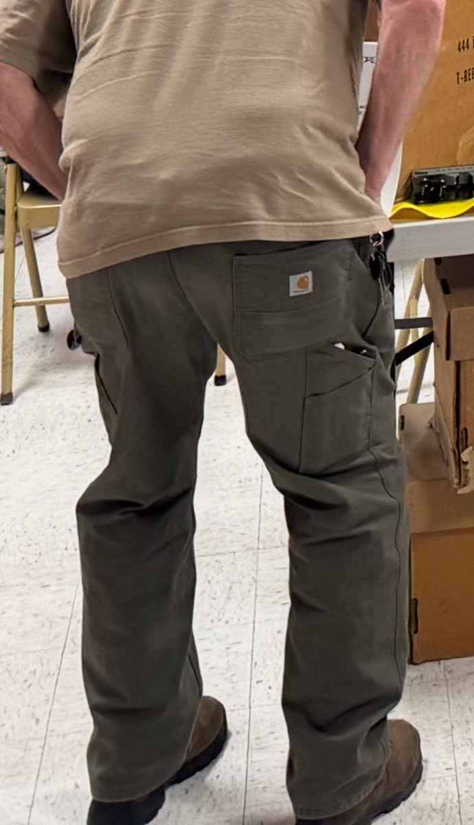 A worker seen from behind wearing green Carhartt carpenter pants with utility pockets, standing among cardboard boxes