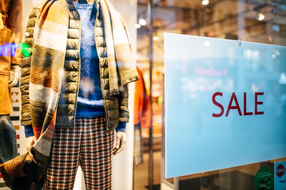 A shop-window mannequin in a plaid scarf, quilted vest and checked trousers beside a pale blue SALE sign