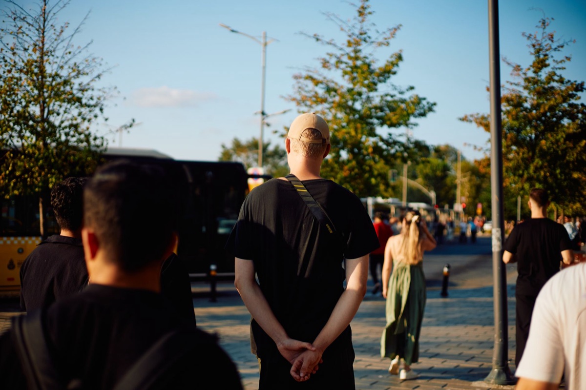 A crowd of pedestrians in short sleeves walking down a sunlit city street lined with green trees
